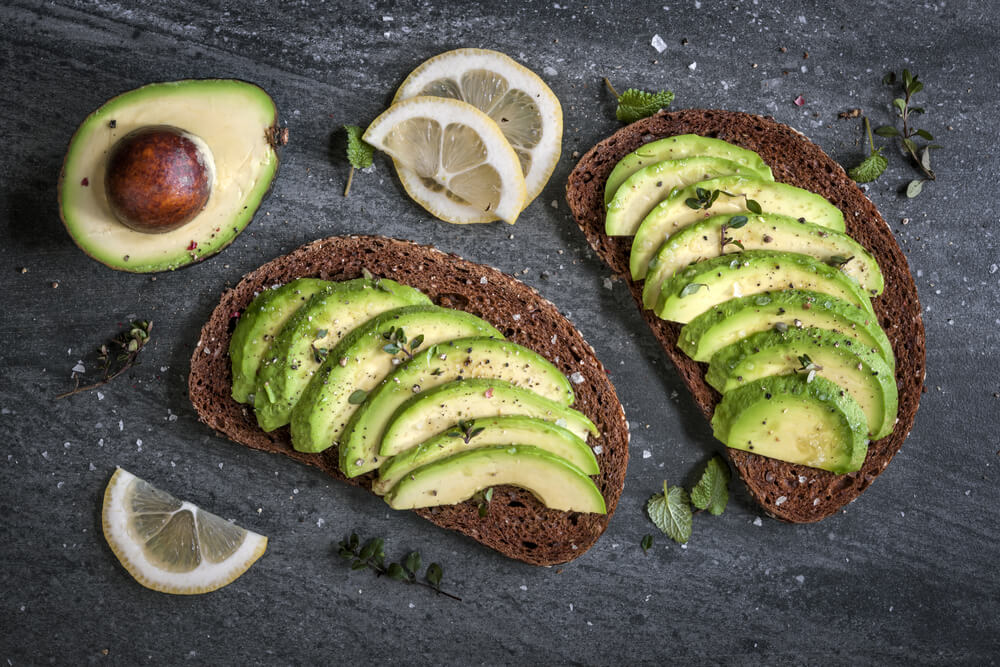 Two slices of dark bread, with sliced avocado on each. A half of an avocado is on the upper left portion of the photo, pit still in. Sliced portions of lemon are scattered around the bread slices.